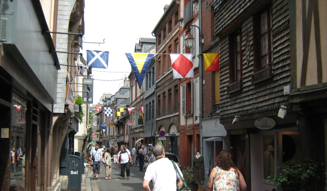 a narrow and old street with overhanging buildings and bunting, busy and bustling with shoppers